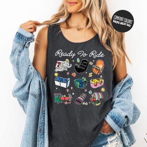 A woman wearing a gray tank top with a graphic design of various cartoon-style objects and text, including the phrase "Ready to Ride".
