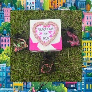 May include: A white box with pink starburst designs and a heart-shaped frame that reads "BRACELETS by BEN." The box is surrounded by small black drawstring bags containing bracelets, all set on a bed of green grass.