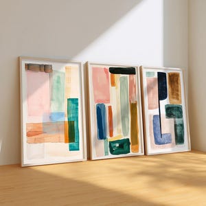 May include: Three framed abstract art prints. Each print features colorful geometric shapes in shades of pink, blue, green, and brown. The artwork is displayed against a white wall and rests on a wooden floor.