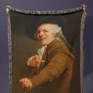 May include: A woven throw blanket featuring a portrait of a man in a brown coat and hat, pointing with a cane in his hand. The blanket has a fringed edge and is held up against a dark blue wall.