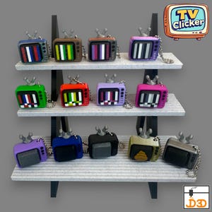 Puede incluir: Una exhibición de llaveros de televisión en miniatura de colores. Cada llavero es un pequeño televisor de estilo retro con una pantalla que muestra una imagen de prueba. Los llaveros son de varios colores, incluyendo azul, verde, rojo y morado, y se exhiben en una estantería de varios niveles.