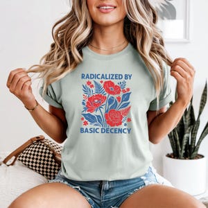 May include: A pale green t-shirt with a floral design of red flowers and blue leaves. The text "RADICALIZED BY BASIC DECENCY" is printed above and below the floral arrangement. The shirt is made of a soft material.
