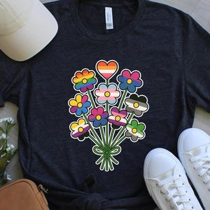 Może przedstawiać: Ciemnoszara koszulka z bukietem kwiatów z różnymi wzorami flag LGBTQ+. Kwiaty są w tęczowych kolorach, z kwiatem w kształcie serca na górze. Koszulka jest połączona z białymi trampkami i białą czapką.
