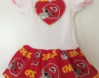 May include: White baby onesie with a red heart featuring a football helmet and the words "KC Chiefs". The onesie has a red and yellow ruffled skirt with a repeated pattern of football helmets and the words "Chiefs".