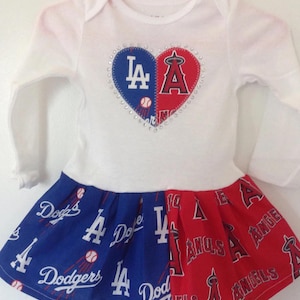 May include: A white baby dress with long sleeves. The bodice has a heart design with the logos of the Los Angeles Dodgers and the Los Angeles Angels. The skirt is split, with blue Dodgers fabric and red Angels fabric.