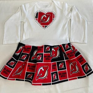 May include: White long-sleeved onesie with a red and black heart-shaped New Jersey Devils logo with rhinestones. The onesie is paired with a black and red skirt with a repeating pattern of the New Jersey Devils logo.