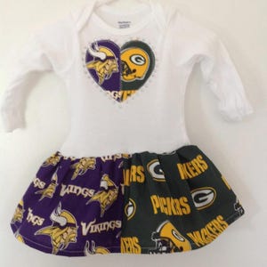 May include: White baby onesie with a heart-shaped applique featuring the logos of the Minnesota Vikings and Green Bay Packers. The onesie has a purple and green skirt with the logos of both teams.