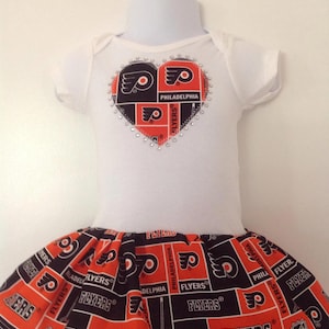 May include: A white baby onesie with a heart-shaped design featuring the Philadelphia Flyers logo and rhinestones. The onesie is paired with a black and orange skirt with a repeating pattern of the Flyers logo.
