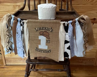 May include: A rustic high chair decorated for a first birthday. A small white cake with a single candle sits on the tray. A banner reads "Lorenzo's 1st Birthday" with a cowboy hat graphic. The banner is adorned with burlap and fabric strips.