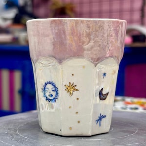 Op de afbeelding: Een witte keramische beker met een gefacetteerde, zeshoekige vorm en een parelmoerachtige, iriserende afwerking. De buitenkant is versierd met kleine, handgeschilderde hemelse motieven, waaronder een blauwe zon met een gezicht, een gouden ster en een maansikkel.