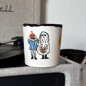 May include: A white ceramic mug featuring a hand-painted illustration of two figures in Halloween costumes. One figure wears a blue and white striped outfit with a pumpkin head, while the other is dressed as a ghost holding a small pumpkin bucket.