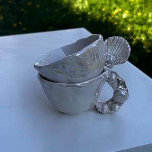 Könnte beinhalten: Zwei weiße Keramik-Teetassen, die übereinander gestapelt sind. Die obere Tasse hat einen unregelmäßigen Rand und eine irisierende Glasur. Die untere Tasse hat einen Griff in Form von Muscheln. Auf weißer Oberfläche mit verschwommenem grünem Hintergrund.
