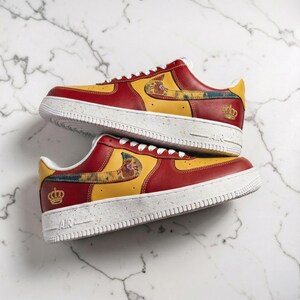 Puede incluir: Zapatillas rojas y amarillas con suelas blancas. Las zapatillas presentan un diseño de la bandera española en el lateral y un emblema de corona dorada. Las suelas tienen un patrón moteado y los cordones son blancos.