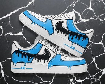 May include: Pair of white sneakers with blue and black paint drip design. The shoes have a white sole, blue accents, and a black swoosh logo. The word "AIR" is printed on the side.
