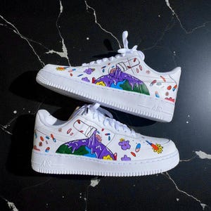 Puede incluir: Zapatillas blancas con ilustraciones coloridas personalizadas. Los zapatos presentan un diseño pintado con una montaña púrpura, una botella y varios elementos florales y abstractos en rojo, amarillo y azul. Los zapatos tienen cordones y suelas blancas.