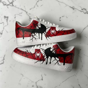 Puede incluir: Par de zapatillas blancas con diseño de telaraña roja y negra. Las zapatillas presentan un logotipo negro, un emblema de araña blanco y detalles de pintura negra goteando. Las suelas son blancas.