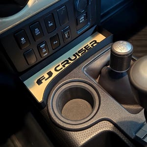 May include: Interior view of a vehicle's center console. The console features a black and beige color scheme, with the text "FJ CRUISER" in gold lettering. Visible are cup holders, gear shift, and various control buttons.