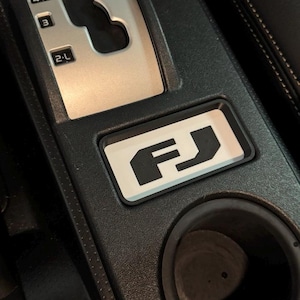 May include: A black car interior with a silver gear shifter and a white FJ emblem on a black panel. The panel also has a cup holder.