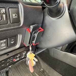 May include: A black and red key holder installed in a vehicle's dashboard, holding a car key and a set of keys. The key holder is mounted near the steering wheel and dashboard controls.