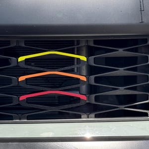 May include: A black car grille with a honeycomb pattern. Three horizontal stripes are placed on the grille, in yellow, orange, and red.