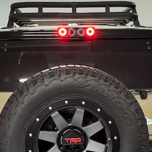 May include: Tan and black SUV with a spare tire and a roof rack. The rear window has a third brake light. The spare tire has a black rim with a red TRD logo. The vehicle's taillight is illuminated.