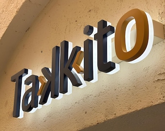 May include: A three-dimensional wall-mounted sign displaying the word Taskito in dark blue and mustard yellow lettering. The letters feature a white backlit glow, creating a halo effect against the textured beige wall surface.