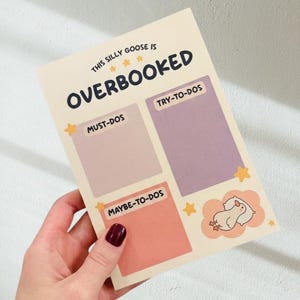 以下が含まれることがあります： 上部に「THIS SILLY GOOSE IS OVERBOOKED」というテキストが書かれたクリーム色のメモ帳。MUST-DOS、TRY-TO-DOS、MAYBE-TO-DOSとラベル付けされた薄いピンク、紫、オレンジの3つの長方形セクションと、小さなガチョウのイラストが描かれています。