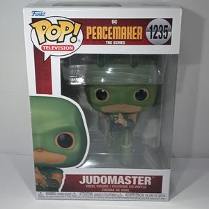 Może przedstawiać: Figurka winylowa Funko Pop! Judomaster z serialu DC Peacemaker, prezentowana w przezroczystej gablocie. Figurka jest zielono-brązowa z dużymi czarnymi oczami. Pudełko zawiera logo Funko Pop! i Peacemaker.