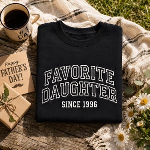 May include: Black sweatshirt with the words "FAVORITE DAUGHTER" in white, arched lettering, and "SINCE 1996" below. A coffee mug with "#1 DAD" is visible, along with a Father's Day card and a small gift box.