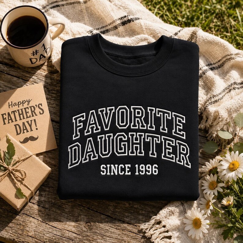 May include: Black sweatshirt with the words "FAVORITE DAUGHTER" in white, arched lettering, and "SINCE 1996" below. A coffee mug with "#1 DAD" is visible, along with a Father's Day card and a small gift box.