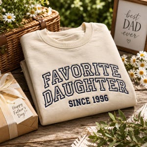 May include: Cream-colored sweatshirt with the embroidered text "FAVORITE DAUGHTER SINCE 1996" in navy blue. A gift box with a "Happy Father's Day!" tag and a framed sign that says "best DAD ever" are also in the image.