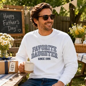 May include: A white sweatshirt with the text "FAVORITE DAUGHTER SINCE 1996" in navy blue lettering. The person is wearing sunglasses and holding a mug. A sign in the background says "Happy Father's Day!"