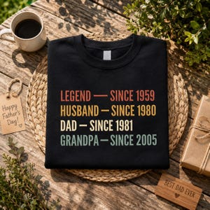 May include: Black sweatshirt with the text "LEGEND - SINCE 1959, HUSBAND - SINCE 1980, DAD - SINCE 1981, GRANDPA - SINCE 2005" in retro colours. A coffee mug, a "Happy Father's Day" tag, and a "Best Dad Ever" sign are also visible.