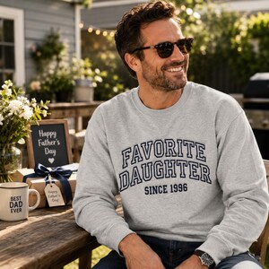 May include: A light gray sweatshirt with the text "FAVORITE DAUGHTER SINCE 1996" in navy blue. A mug that says "BEST DAD EVER" is on a wooden table. A sign says "Happy Father's Day".