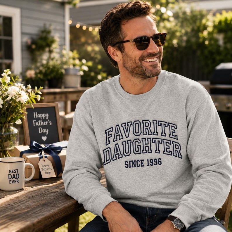 May include: A light gray sweatshirt with the text "FAVORITE DAUGHTER SINCE 1996" in navy blue. A mug that says "BEST DAD EVER" is on a wooden table. A sign says "Happy Father's Day".