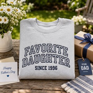 May include: A light gray sweatshirt with the words "FAVORITE DAUGHTER SINCE 1996" embroidered in navy blue. A gift box with a blue ribbon and a "FOR DAD" tag are next to the sweatshirt. A card that says "Happy Father's Day" is also present.