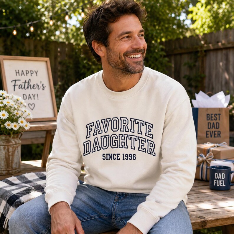 May include: Cream-colored sweatshirt with the text "FAVORITE DAUGHTER SINCE 1996" in navy blue. The image also includes a sign that says "Happy Father's Day!" and a gift bag that says "Best Dad Ever."