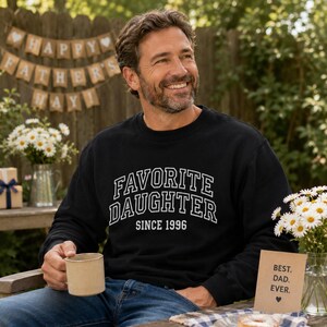 May include: A black sweatshirt with the text "FAVORITE DAUGHTER SINCE 1996" in white lettering. The image shows a man holding a mug, with a "Happy Father's Day" banner in the background. A card that says "Best. Dad. Ever." is also visible.