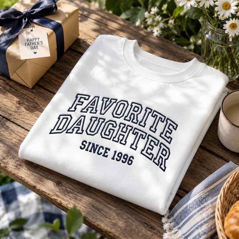 May include: White sweatshirt with the text "FAVORITE DAUGHTER SINCE 1996" in navy blue. A gift box with a navy blue ribbon and a "Happy Father's Day" tag is visible. The items are on a wooden surface with flowers and a cup.