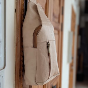 May include: A beige leather sling bag with a zippered main compartment and an exterior pocket. The bag has a long strap and is hanging on a wooden door. The bag's dimensions are not visible.