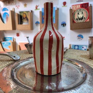 Pode incluir: Um vaso de cerâmica branca com listras verticais vermelhas. O vaso tem um pescoço estreito e um corpo arredondado.