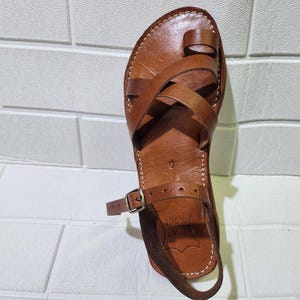 Peut inclure: Sandales en cuir marron avec des lanières croisées et une boucle d'orteil. Les sandales ont une fermeture à boucle et le texte "100%  CUERO" est visible sur la semelle.