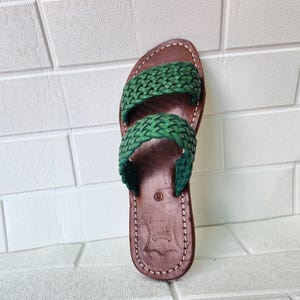 Puede incluir: Sandalia de cuero marrón con dos correas verdes trenzadas. La sandalia tiene una suela cosida y una plantilla de cuero texturizado. El diseño es sencillo y adecuado para uso informal. La sandalia se muestra contra una pared de ladrillos blancos.