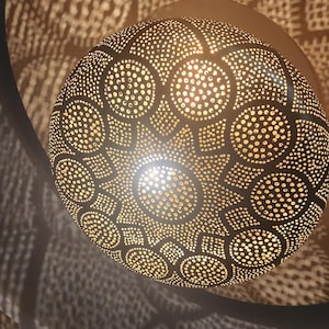 May include: A round, gold-colored metal lampshade with a decorative pattern of small circles and larger circles. The lampshade is lit from within, casting a warm glow.