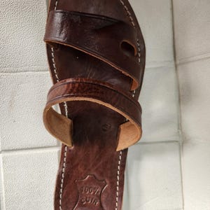 May include: Brown leather sandals with a single strap across the top and two straps around the ankle. The sandals have a 100% cuir label on the bottom.