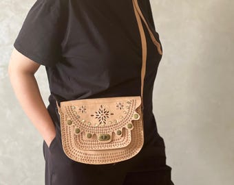 以下が含まれることがあります： 装飾的な花のカットアウトデザインが施された、明るいタン色の革製クロスボディバッグ。 バッグは金属製の留め金が付いたフラップ開閉式で、長くて調節可能なストラップが付いています。 バッグには、小さくて丸い金属スタッドが飾られています。