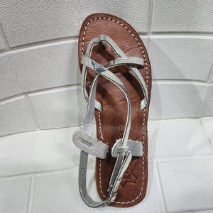 Puede incluir: Sandalia de cuero marrón con costuras blancas y correas grises. La sandalia tiene un diseño tipo tanga con múltiples correas que cruzan el pie y alrededor del tobillo. La suela es de cuero marrón.