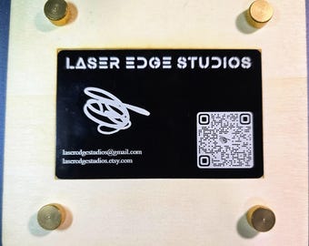May include: A wooden plaque with a black background and white text that reads "LASER EDGE STUDIOS". The plaque has a QR code and contact information for the business. The plaque is attached to a wooden base with four brass screws.