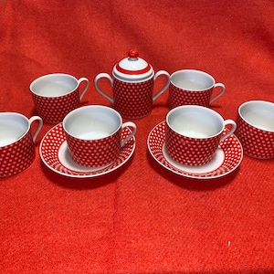 Puede incluir: Un juego de té de lunares rojos y blancos, que incluye seis tazas, platillos y una tetera con tapa. Las tazas y la tetera tienen un interior blanco. El juego está dispuesto sobre una superficie roja.