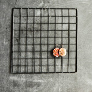 May include: A pair of round, orange and white patterned earrings hanging on a black wire rack.